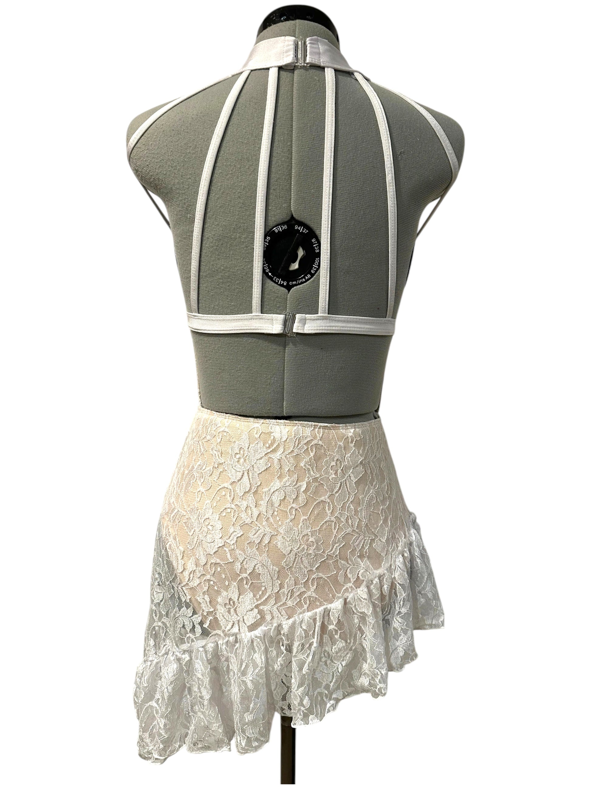 Back view of the white lace Demi dance costume base featuring satin strap detailing, open back design, illusion high neckline, and asymmetrical lace ruffle skirt.