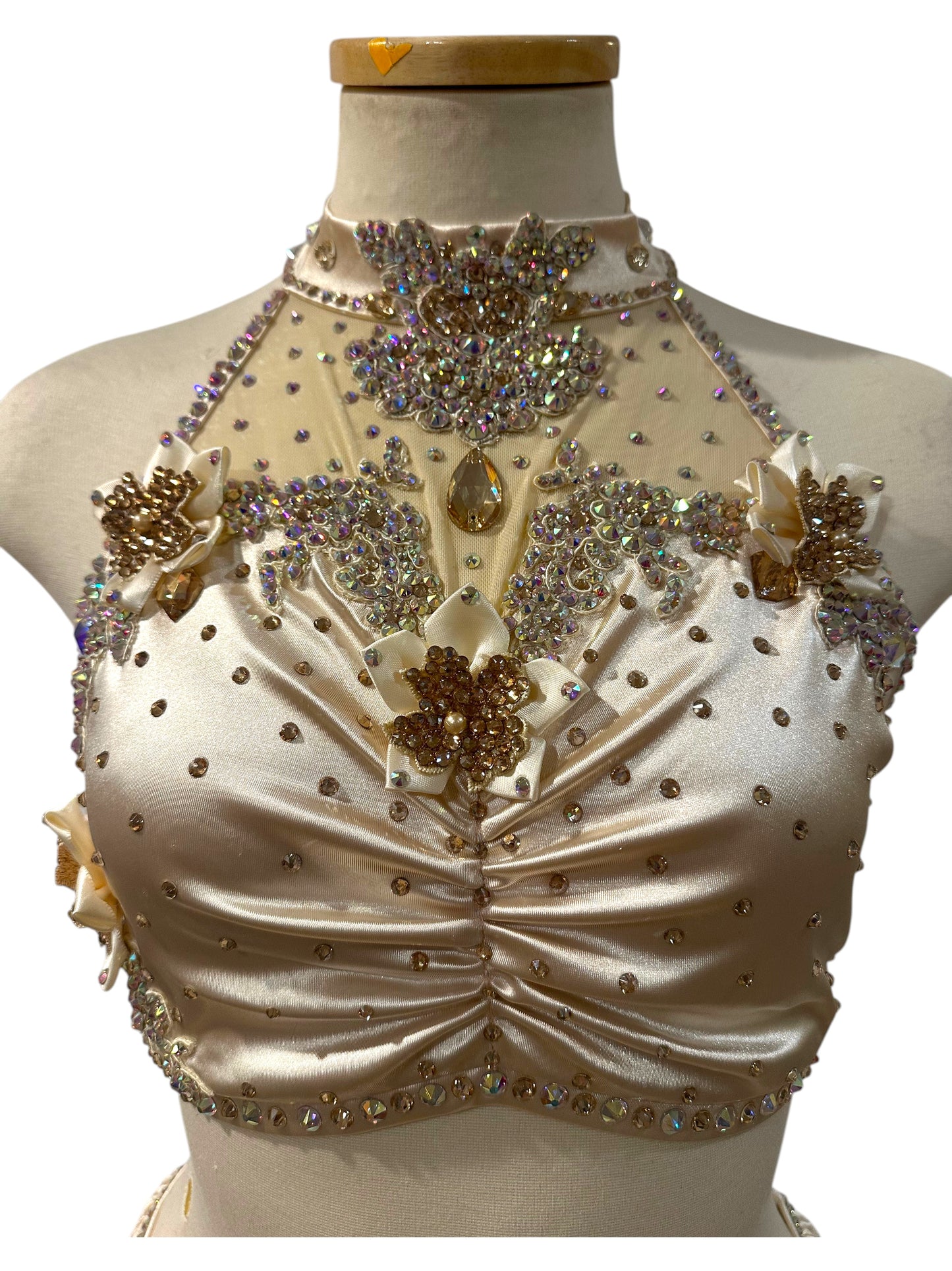 Close-up of the Aubrey Ready-to-Ship dance costume featuring almond white satin, hand-placed crystal appliqués, dimensional floral accents, illusion mesh, and a rhinestone halter neckline.
